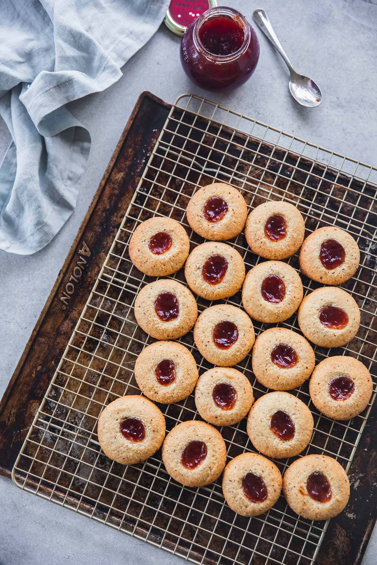 Freshly baked Frangipane Jam Drop Biscuits cooling on a wire rack with a jam bottle, spoon and tea towel to the side.