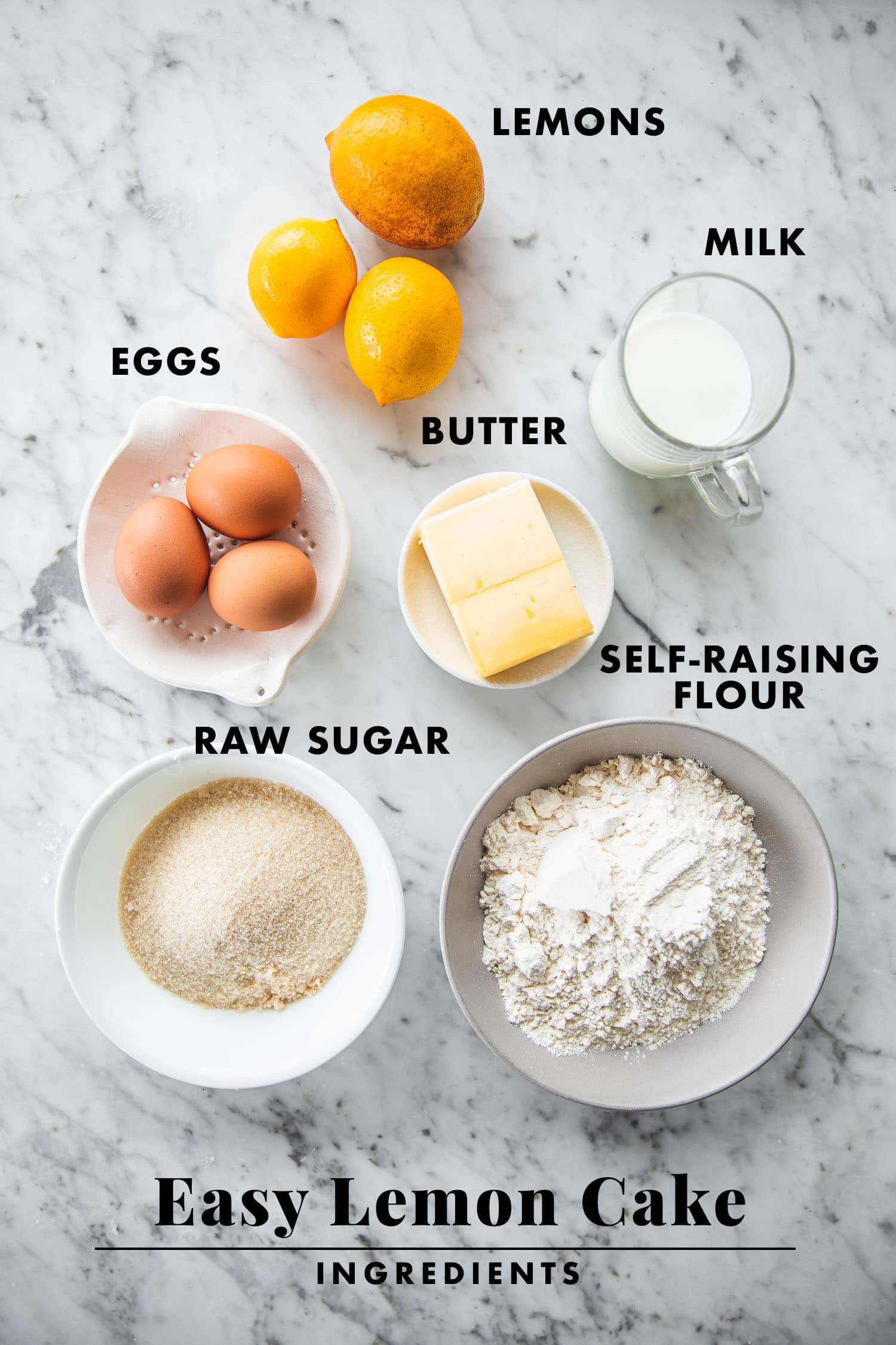 Ingredients for lemon cake set out on a table - flour, eggs, sugar, butter, milk and lemons.