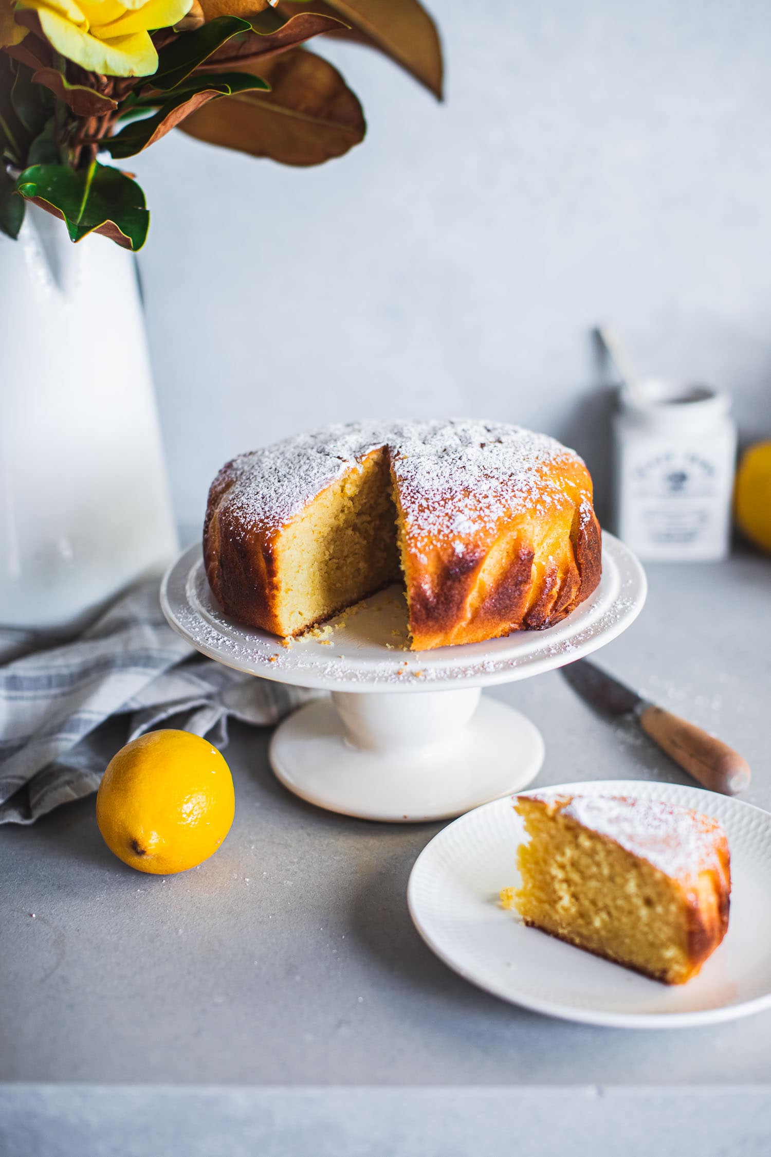 A whole baked lemon cake on a cake stand, a lemon cake slice served on a plate, lemons and tea towel on a kitchen counter.