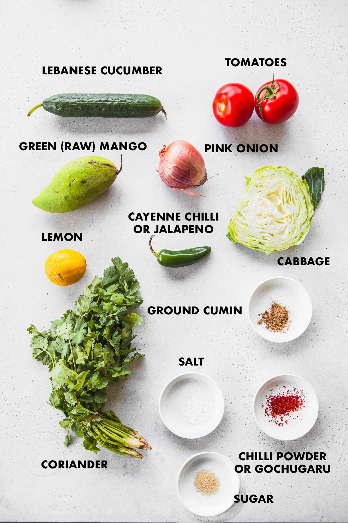 Kachumber Salad ingredients labelled - cucumber, tomatoes, raw green mango, pink onion, cabbage, green chilli, coriander, lemon, cumin, chilli powder, salt and sugar.