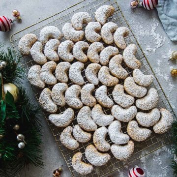 Kourabiedes - Greek Almond Crescent Cookies dusted with icing sugar on a wire rack surrounded by Christmas wreath and ornaments.