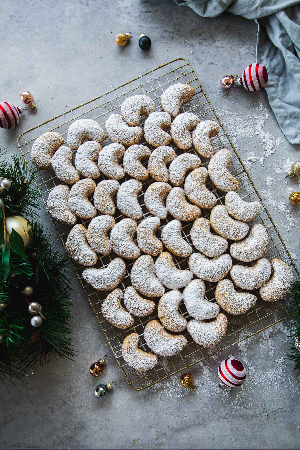 Kourabiedes - Greek Almond Crescent Cookies dusted with icing sugar on a wire rack surrounded by Christmas wreath and ornaments.