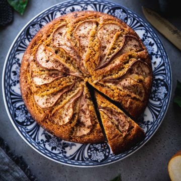 Spiced Pear Cake sliced and served on a floral blue plate with tea towels, cake knife and pears on the table.