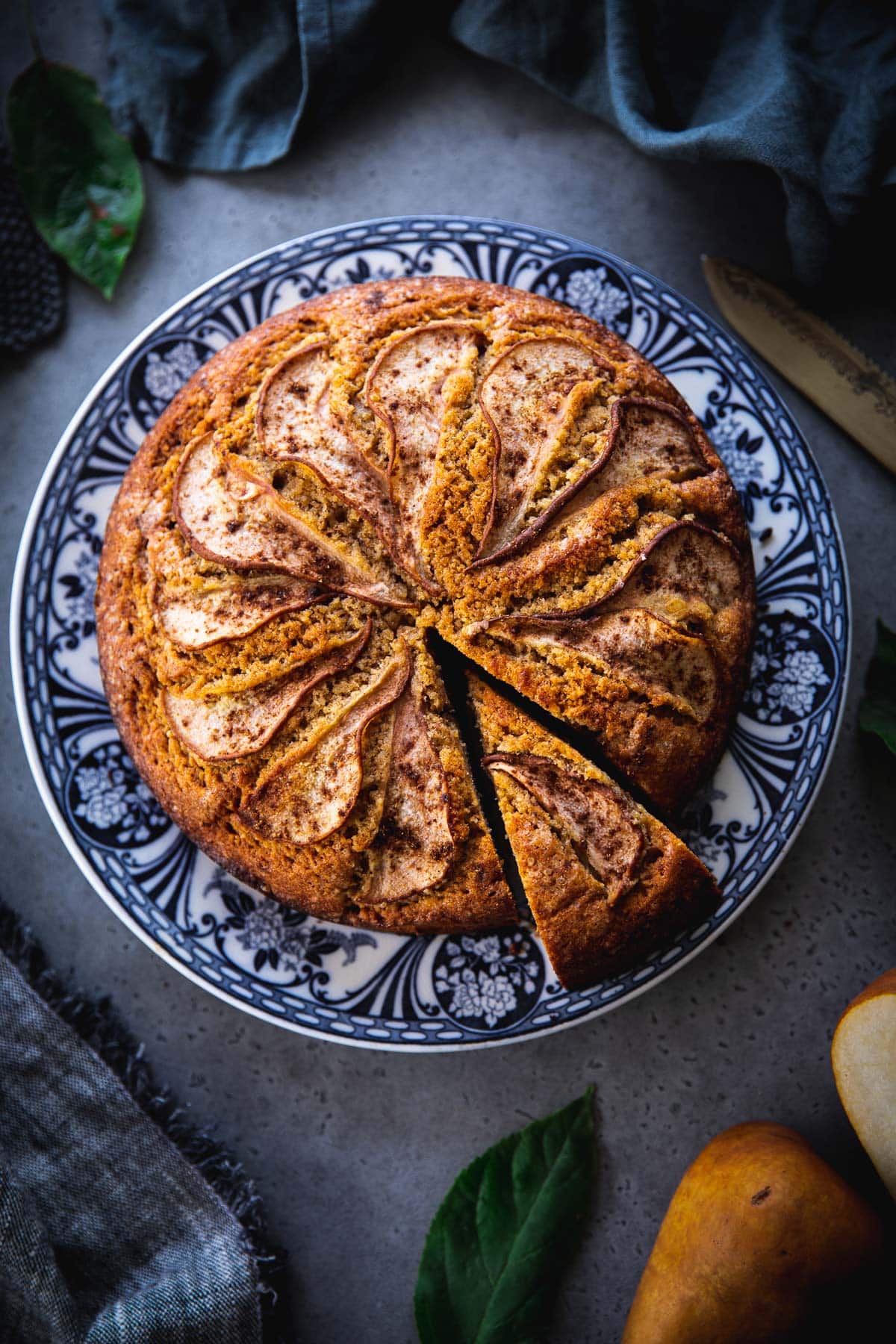 Spiced Pear Cake sliced and served on a floral blue plate with tea towels, cake knife and pears on the table.