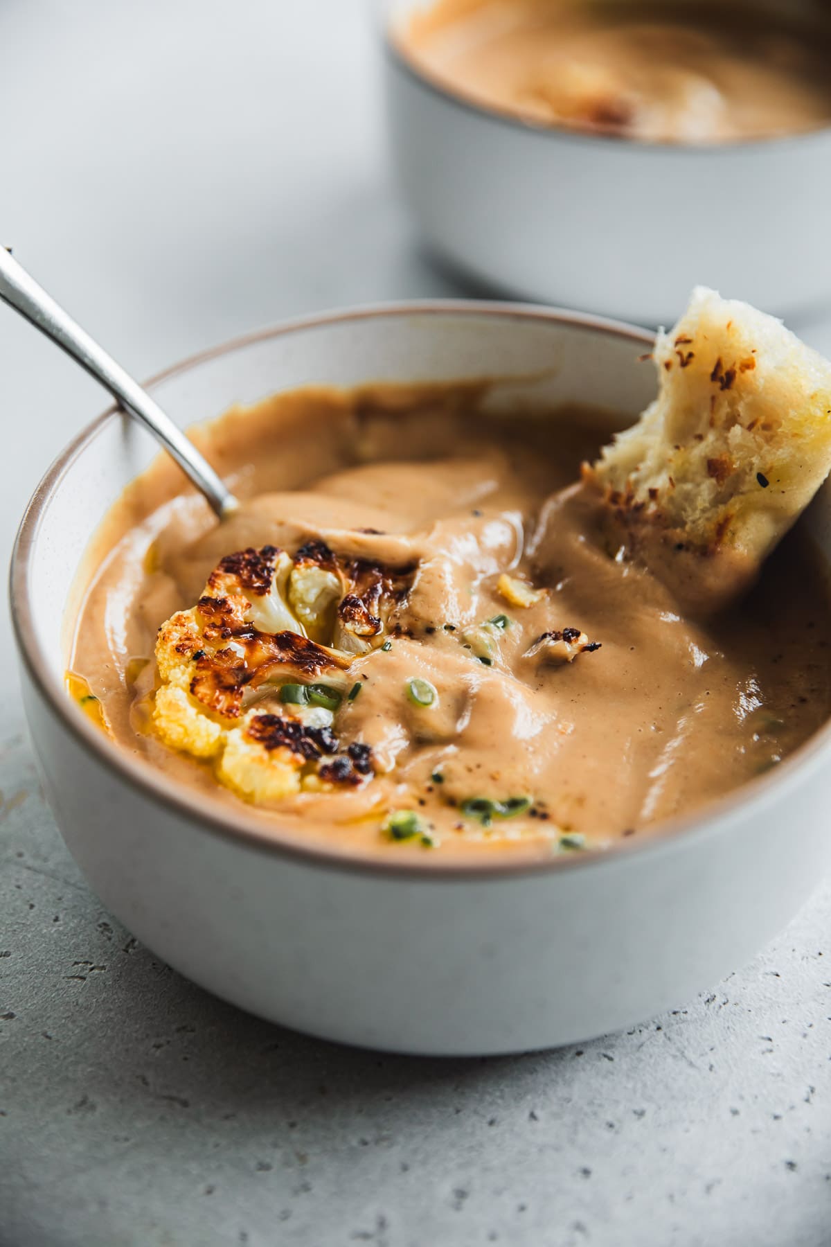 Thick, creamy roasted cauliflower soup in a bowl with a spoon and toasted bread stick.