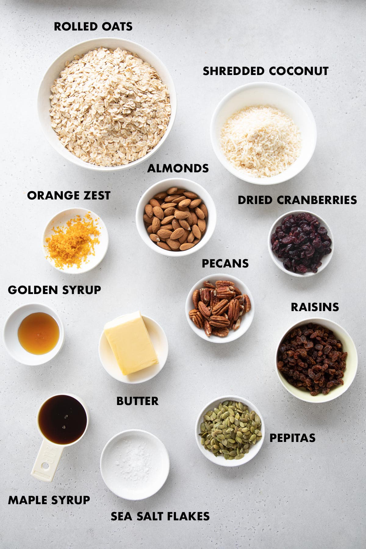 Homemade Healthy Granola Ingredients measured in bowls and labeled - rolled oats, shredded coconut, almonds, pecans, dried cranberries, raisins, golden syrup, maple syrup, orange zest, butter, pepitas and salt.