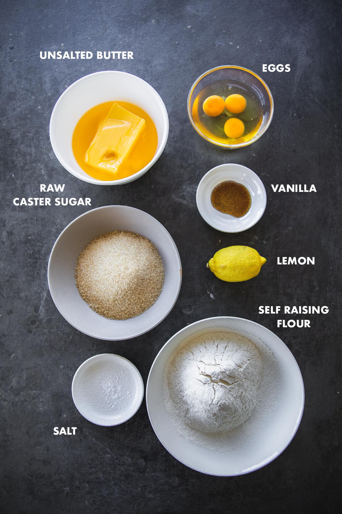 Ingredients for lemon madeira cake measured out in bowls and labeled - self raising flour, vanilla, lemon, caster sugar, eggs and unsalted butter.
