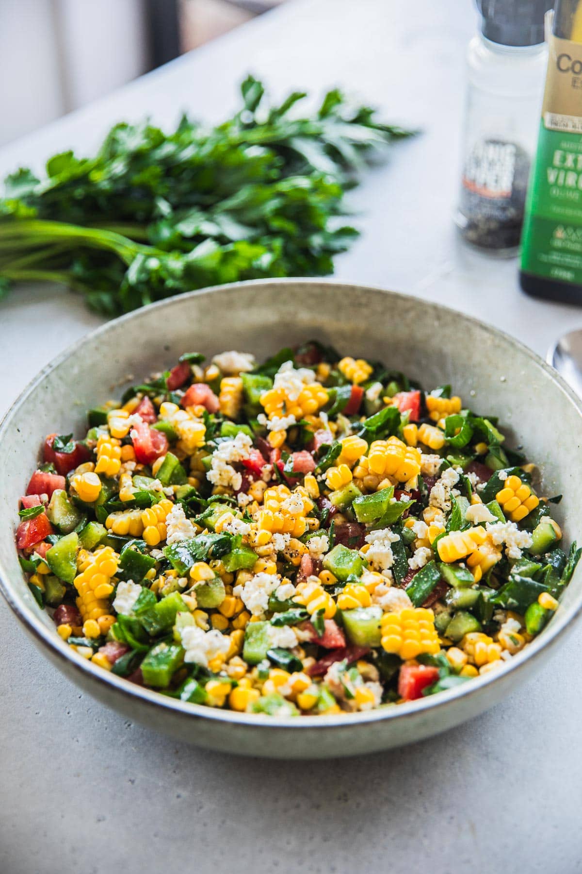 Feta Corn Salad in a bowl on the table with a parsley bunch and olive oil bottle.