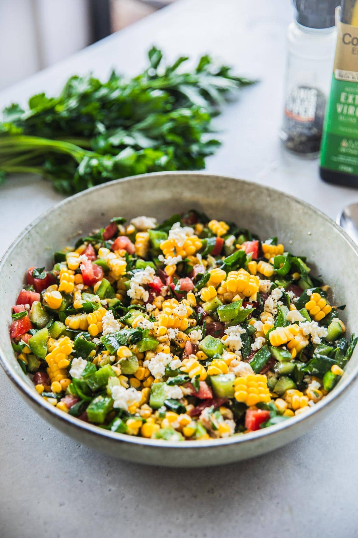 Feta Corn Salad in a bowl on the table with a parsley bunch and olive oil bottle.