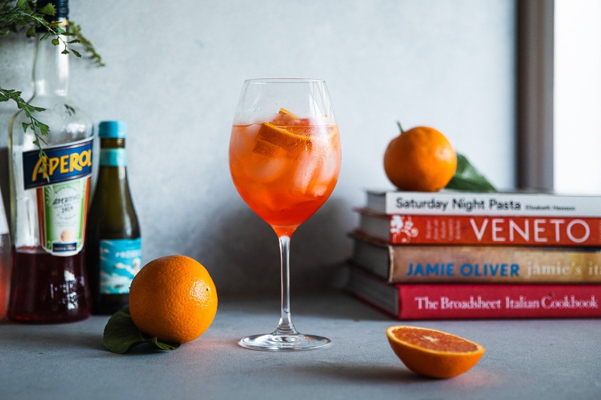 Aperol Spritz cocktail in a glass on a counter with bottles of Aperol and Prosecco, an orange and a stack of cookbooks.