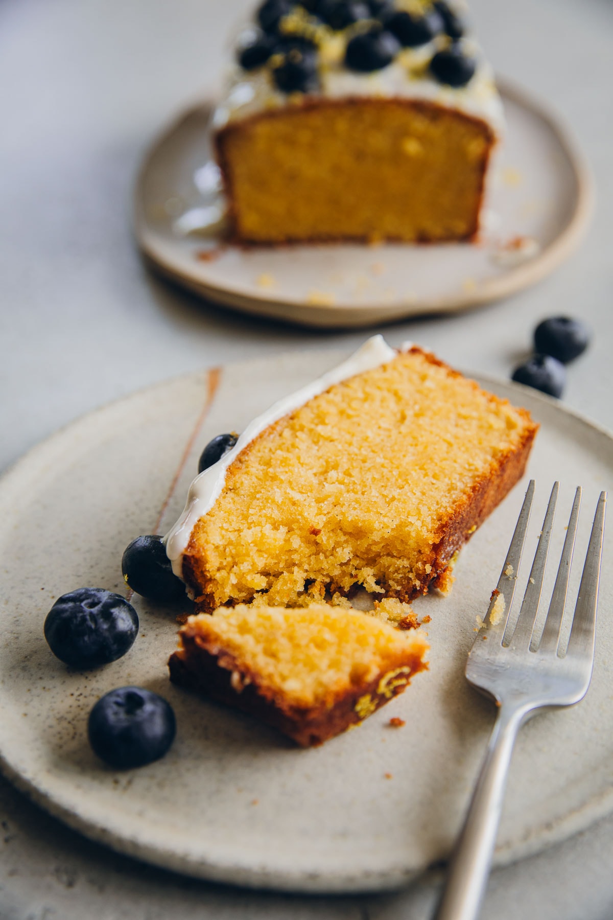 A slice of Blueberry Lemon Loaf Cake With Yoghurt Icing and fresh blueberries on a plate with a fork.