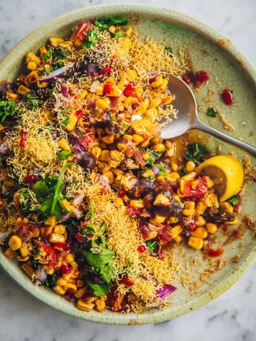 Corn Chaat served in a green plate topped with sev, onion, coriander, sweet date chutney and lemon wedge.