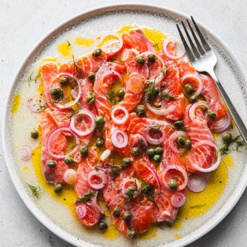 Salmon Crudo served on a plate - slices of sashimi grade raw salmon topped with a lemon olive oil dressing, onion, cxapers and dill.